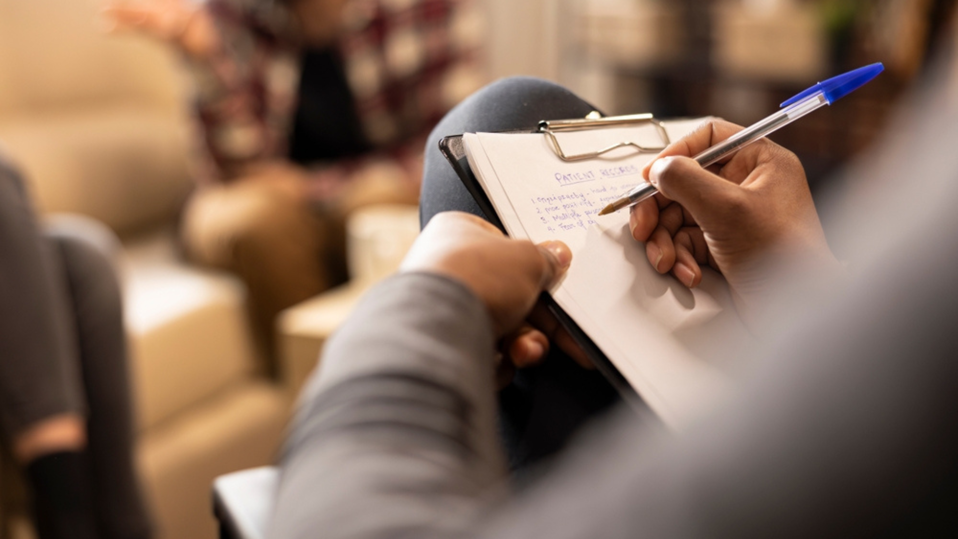Close up of African American hands writing with pen on paper, documenting symptoms. A patient sitting on a sofa is blurred in the background.