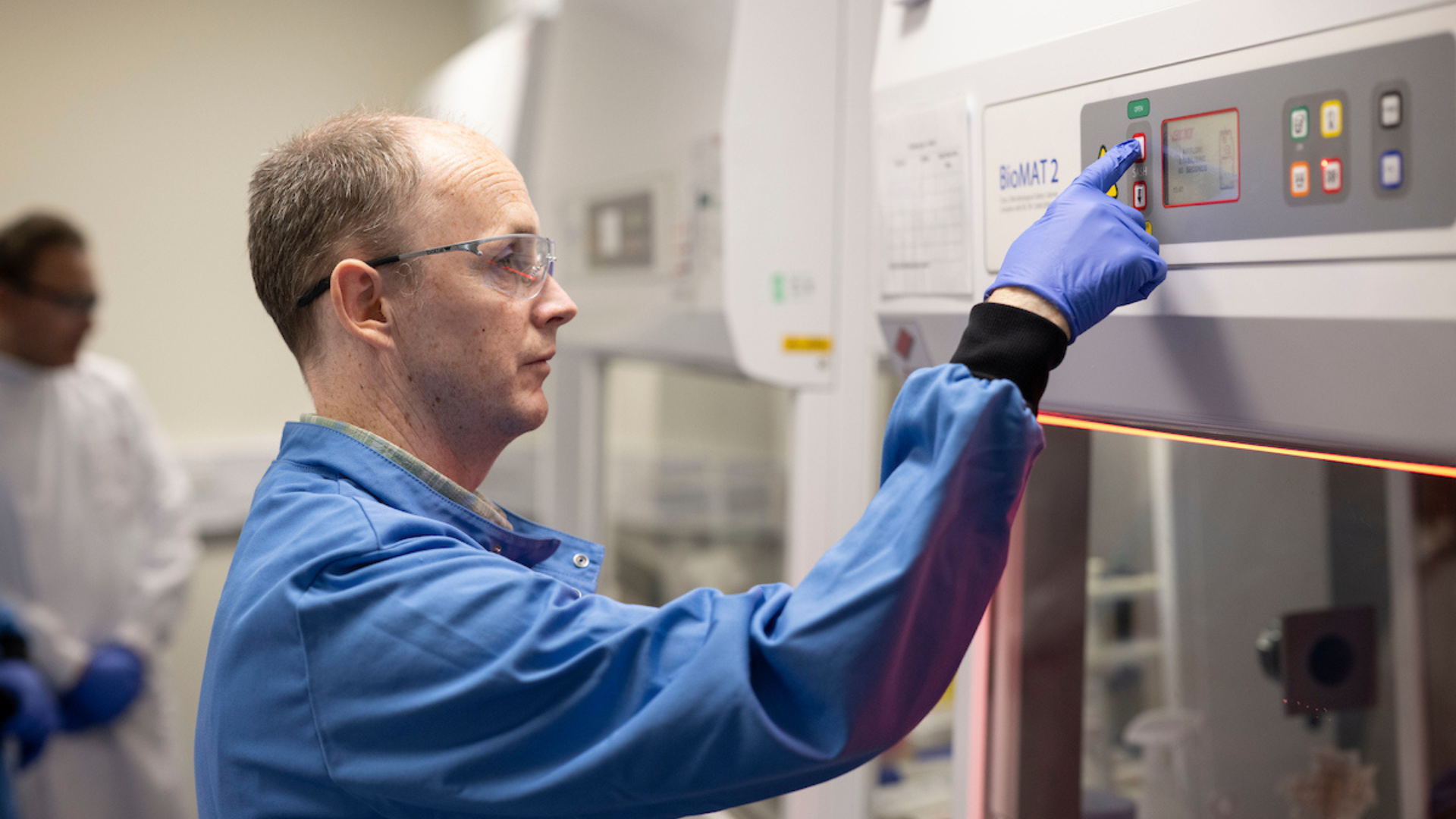 A man in a labcoat stands at a biological safety cabinet pressing a button. He wears safety goggles and blue medical gloves.