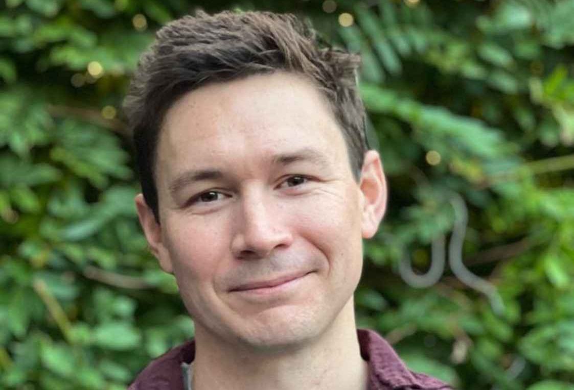 Professional headshot of Sean Willcock, Departmental Lecturer in the History of Art, wearing a red jumper, looking at the camera against a hedge.