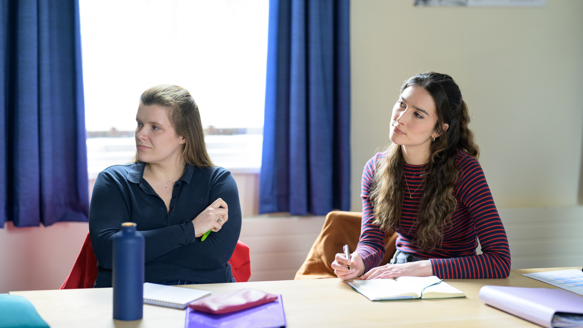 Two women in a classroom sitting at a desk with notebooks in front of them, listening.
