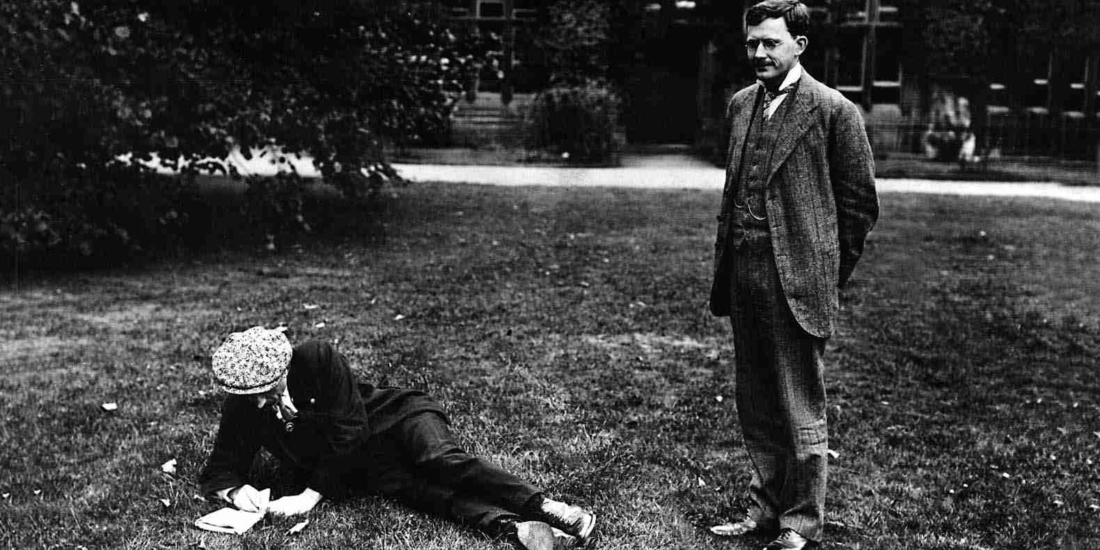 Black and white image of Albert Mansbridge standing on the lawn and another figure lying on the grass making notes.