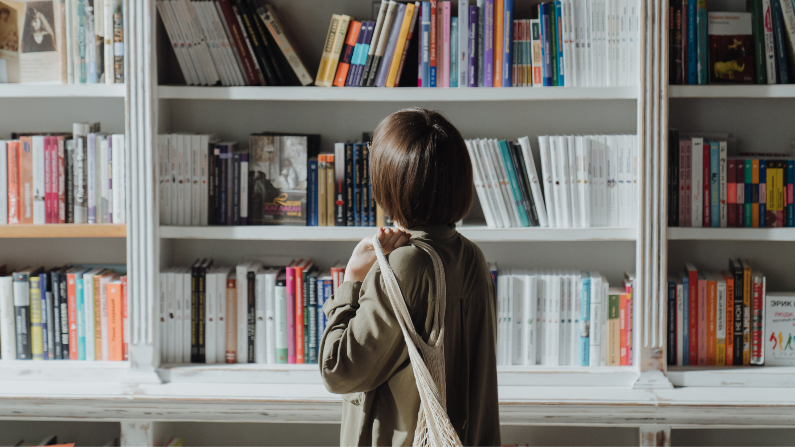 A woman is facing a bookshelf holding a net bag over her shoulder, looking up at the array of books towering above her.