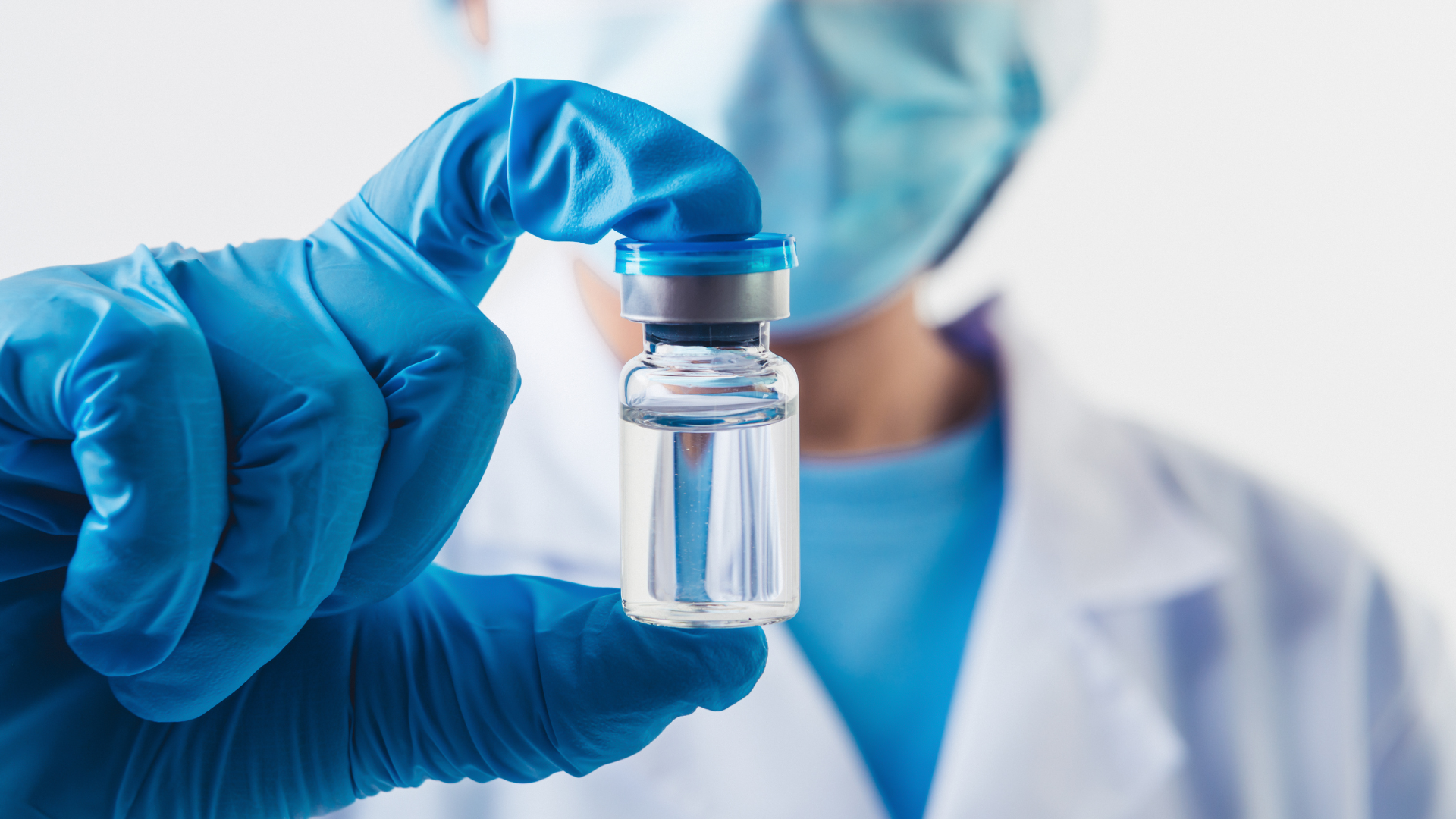 A hand wearing a blue plastic glove holds a vial of clear liquid in the foreground. In the background is a person wearing a mask and medical jacket.