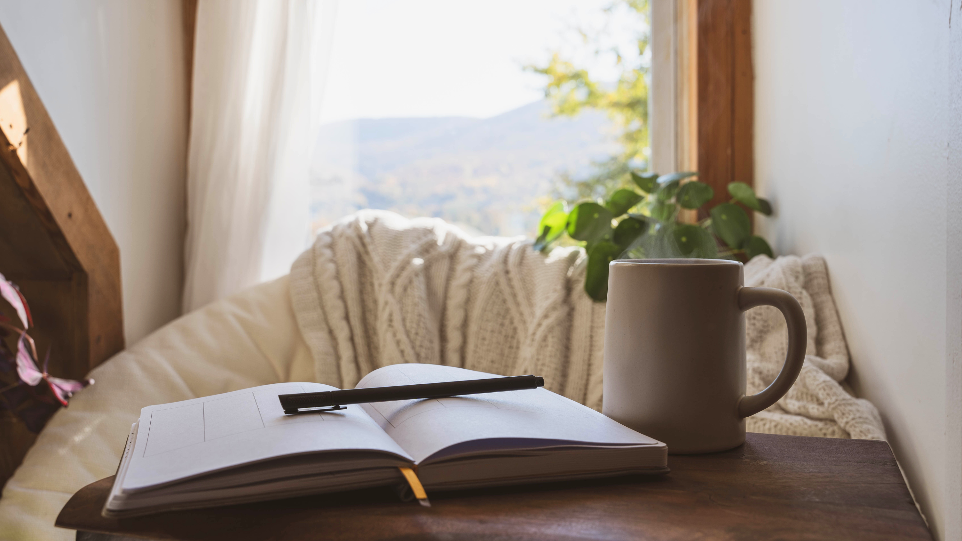 A journal with a pen and a mug with steam are placed on a table, by a comfortable chair with a knit jumper draped on it. In the background is a window overlooking green hills on a bright day, with a houseplant on the sill.