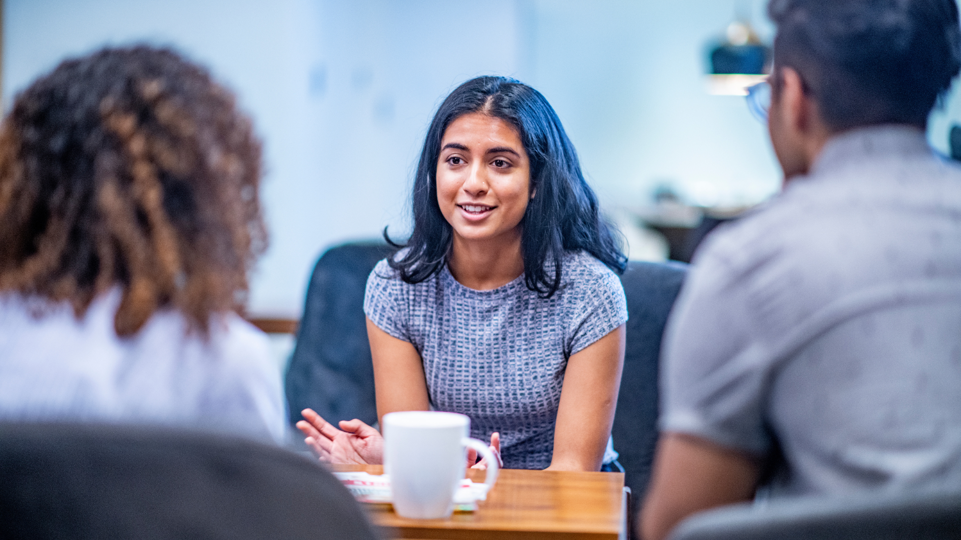 A group of 3 young adults sat around a table, having a discussion and smiling.