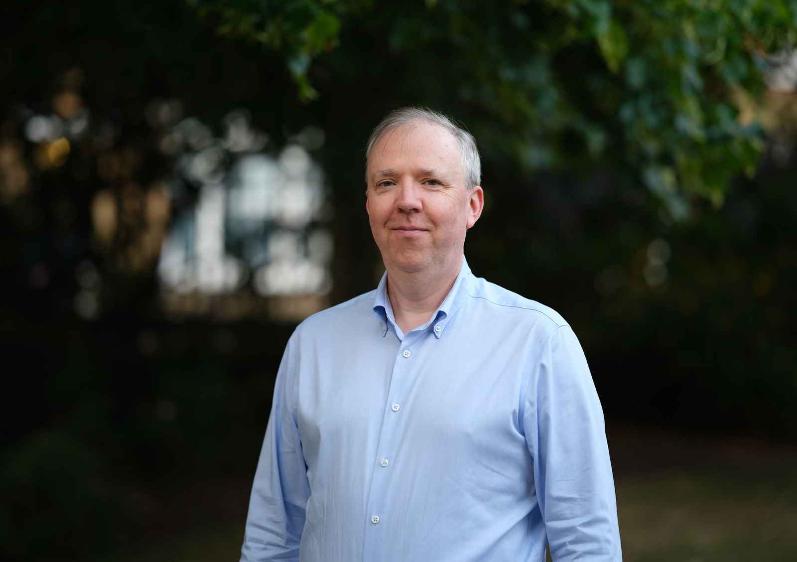 Professional headshot of Joe MacDonagh, Visiting Fellow, wearing a blue shirt, looking at the camera against a blurred background.