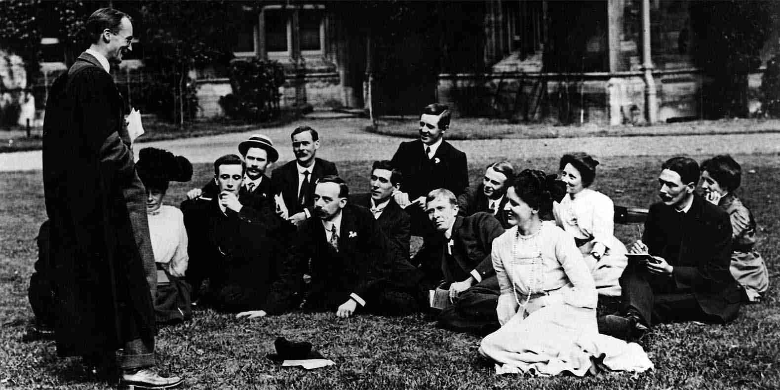 Black and white image of students in Victorian dress sitting on the lawn of an Oxford college, taking notes and listening to their tutor who stands in front of them.